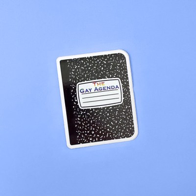 A sticker of a composition book with rainbow text The Gay Agenda as the book title